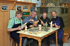 4 Schnitzer sitzen bei ihrem Hobby am Tisch. Im Hintergrund erwärmt ein Kachelofen den Raum.